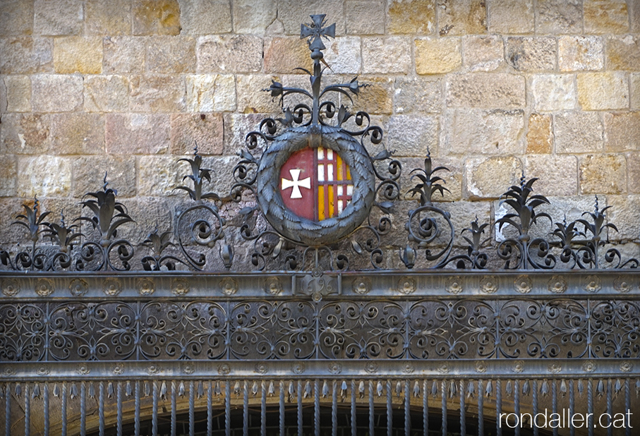 Hospital de la Santa Creu. Reixa de forja a la finestra de l'antiga farmàcia, amb l'escut de la Catedral i del Consell de Cent.