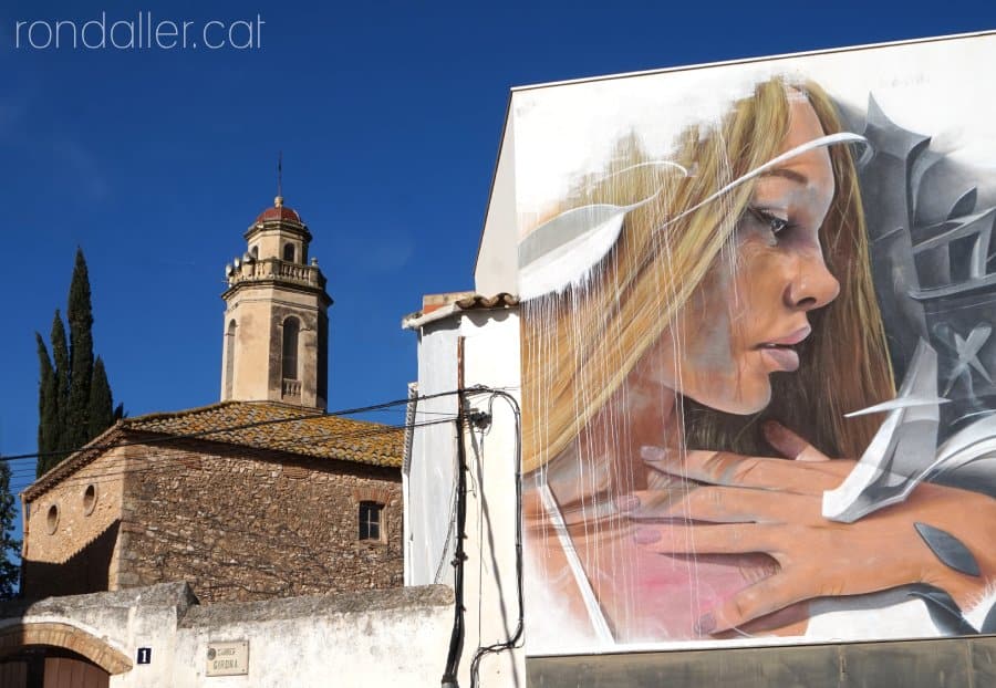 Vista d'un mural pintat i de fons, el campanar de l'església parroquial.